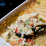 A large black serving spoon scoops a portion of the casserole out of the baking dish. Chicken, peas, and carrots are visible.