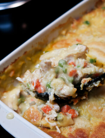 A large black serving spoon scoops a portion of the casserole out of the baking dish. Chicken, peas, and carrots are visible.