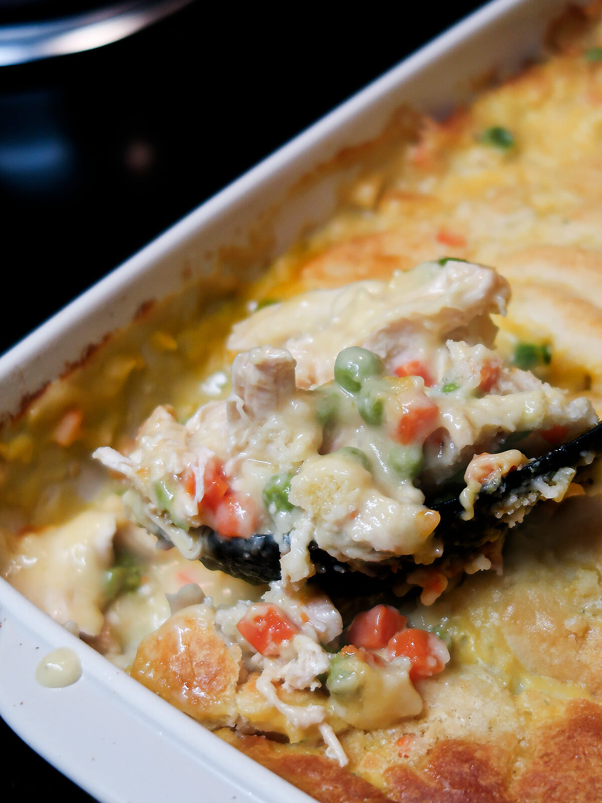 A large black serving spoon scoops a portion of the casserole out of the baking dish. Chicken, peas, and carrots are visible.
